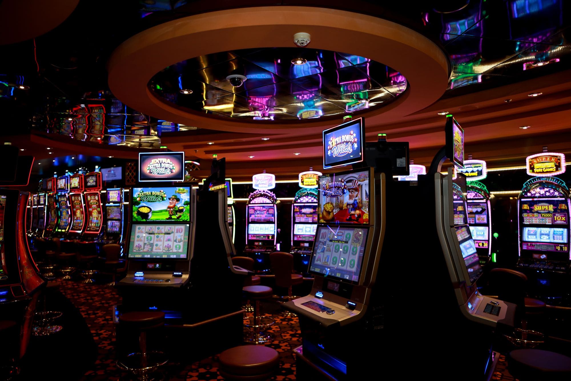 A casino floor at night, rows of slot cabinets lit in pink and blue neon under a mirrored ceiling